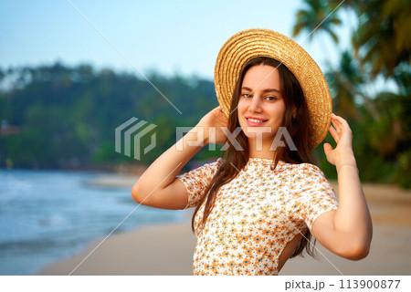 Smiling young woman in summer dress and straw hat stands on tropical beach at sunset. Female traveler enjoys exotic coast, warm sea breeze, palm backdrop. Leisure, vacation vibe by ocean. 113900877