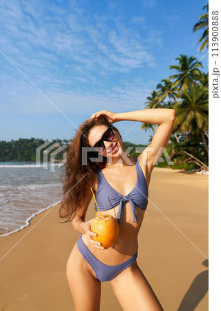 Happy woman in bikini holds fresh coconut on sunny tropical beach. Female enjoys summer by sea with palm trees. Blue sky, clear ocean water, wellness lifestyle. Drinking natural refreshment. Happy woman in bikini holds fresh coconut on sunny tropical beach. Female enjoys summer by sea with palm trees. Blue sky, clear ocean water, wellness lifestyle. Drinking natural refreshment. 113900888