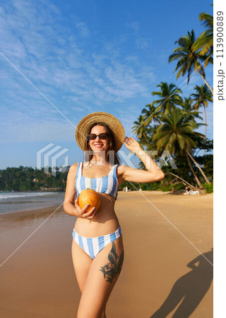 Woman in striped bikini holds fresh coconut on sunny beach. Solo traveler enjoys tropical seaside, healthy drink. Palm trees, blue sky in background. Relaxing, hydration benefits by the sea. Woman in striped bikini holds fresh coconut on sunny beach. Solo traveler enjoys tropical seaside, healthy drink. Palm trees, blue sky in background. Relaxing, hydration benefits by the sea. 113900889