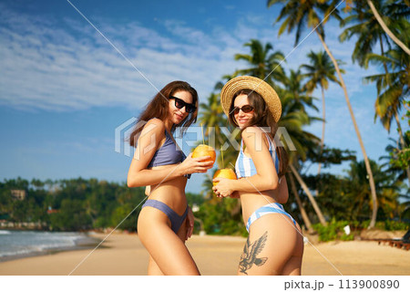 Two women wearing sunglasses and swimwear hold coconuts on sunny beach with palm trees. Friends enjoy tropical vacation, fresh drinks by the blue sea. Travel and leisure with ocean backdrop. Two women wearing sunglasses and swimwear hold coconuts on sunny beach with palm trees. Friends enjoy tropical vacation, fresh drinks by the blue sea. Travel and leisure with ocean backdrop. 113900890