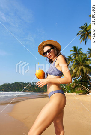 Smiling fit woman in sunglasses and straw hat holds fresh coconut on sunny tropical beach. Bikini-clad traveler enjoys healthy drink, palm trees, blue sky background. Wellness, summer holiday scene. 113900938