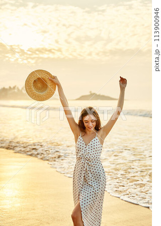 Elegant woman in polka dot dress enjoys sunset at beach, waves gently wash over her bare feet. Arms raised, holding wide-brimmed hat, she smiles, embodying leisure, travel, and seaside freedom. Elegant woman in polka dot dress enjoys sunset at beach, waves gently wash over her bare feet. Arms raised, holding wide-brimmed hat, she smiles, embodying leisure, travel, and seaside freedom. 113900946