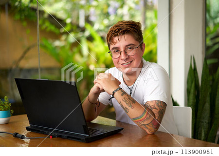 Smiling transgender man works on laptop in modern coworking space. Comfortable inclusive work environment, happy LGBT pro using tech, earphones on desk, indoor plants, natural light. Smiling transgender man works on laptop in modern coworking space. Comfortable inclusive work environment, happy LGBT pro using tech, earphones on desk, indoor plants, natural light. 113900981