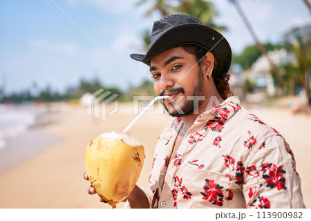 Smiling gay man in floral shirt, hat sips coconut water on sandy beach. Clear blue sky, sunny day, chill vacation vibe. Solo traveler enjoys exotic tropical island, gay-friendly travel theme. Smiling gay man in floral shirt, hat sips coconut water on sandy beach. Clear blue sky, sunny day, chill vacation vibe. Solo traveler enjoys exotic tropical island, gay-friendly travel theme. 113900982