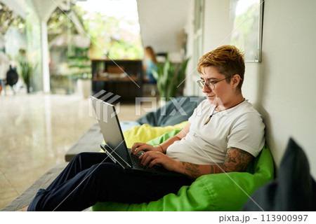 Transgender professional works on laptop, seated on green bean bag, in casual office space. Relaxed environment, light-filled room, modern work life balance, inclusion in workplace highlighted. Transgender professional works on laptop, seated on green bean bag, in casual office space. Relaxed environment, light-filled room, modern work life balance, inclusion in workplace highlighted. 113900997