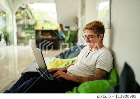 Transgender person lounges on bean bag chair, working on laptop in a light-filled coworking space, reflecting modern work flexibility and inclusion in the professional environment. 113900999