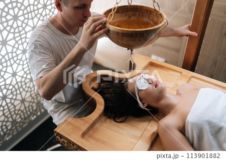 High-angle view of pouring oil on female forehead during exotic Shirodhara treatment, Ayurvedic healing technique . Serene young woman at massage session with aromatic oil dripping on forehead. 113901882