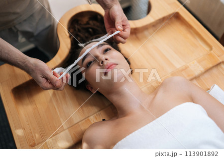 Top view of spa master applying bandage to forehead of young woman client in preparation for an exotic Shirodhara treatment, Ayurvedic healing technique in India salon. Concept of healthy lifestyle. 113901892