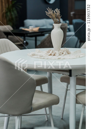 White wooden table complete with white wooden chairs with soft fabric upholstery. Milky porcelain vase on the table 113903732