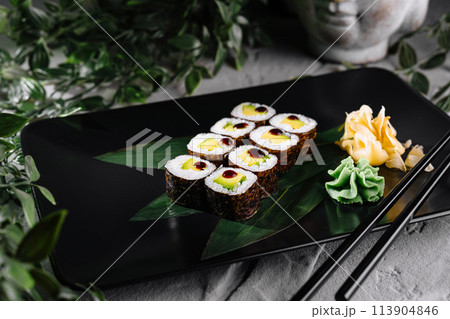 Sushi rolls garnished with wasabi and ginger on a modern black plate, botanical backdrop Sushi rolls garnished with wasabi and ginger on a modern black plate, botanical backdrop 113904846