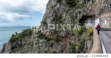 A female tourist walking on the road at the Amalfi Coast, the old historic way is seen overgrown at left side of the road 113906395