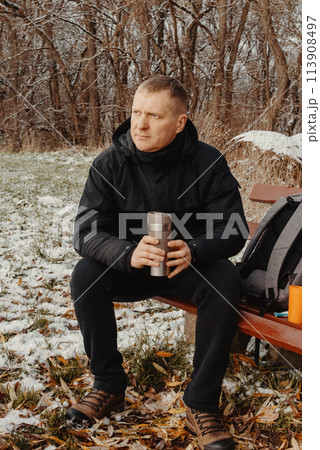 Winter Serenity: 40-Year-Old Man Enjoying Tea on Snow-Covered Bench in Rural Park. Immerse yourself in the tranquil beauty of winter as a 40-year-old man finds solace on a snowy bench in a rural park. 113908497