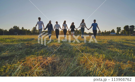 Friends at sunset holding hands walking towards the sun. 113908762