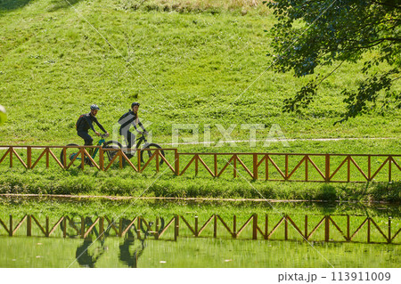 A blissful couple, adorned in professional cycling gear, enjoys a romantic bicycle ride through a park, surrounded by modern natural attractions, radiating love and happiness 113911009