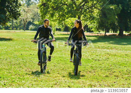 A blissful couple, adorned in professional cycling gear, enjoys a romantic bicycle ride through a park, surrounded by modern natural attractions, radiating love and happiness 113911057