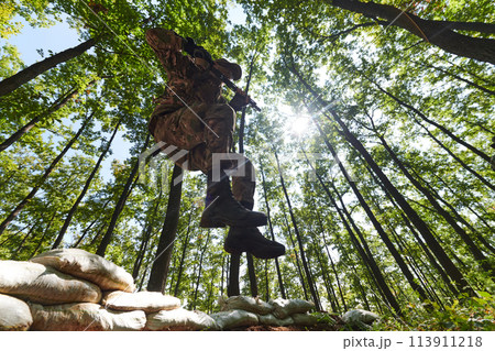 An elite soldier adeptly clears military barriers in the perilous wooded terrain, showcasing tactical skill and agility during specialized training An elite soldier adeptly clears military barriers in the perilous wooded terrain, showcasing tactical skill and agility during specialized training 113911218