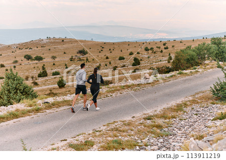 A couple dressed in sportswear runs along a scenic road during an early morning workout, enjoying the fresh air and maintaining a healthy lifestyle A couple dressed in sportswear runs along a scenic road during an early morning workout, enjoying the fresh air and maintaining a healthy lifestyle 113911219