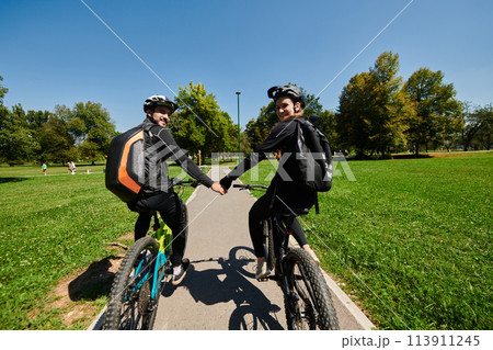 A sweet couple, adorned in cycling gear, rides their bicycles, their hands interlocked in a romantic embrace, capturing the essence of love, adventure, and joy on a sunlit path 113911245