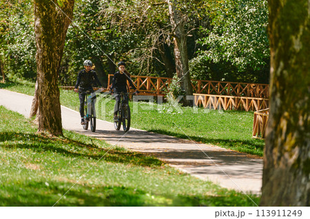 A blissful couple, adorned in professional cycling gear, enjoys a romantic bicycle ride through a park, surrounded by modern natural attractions, radiating love and happiness 113911249