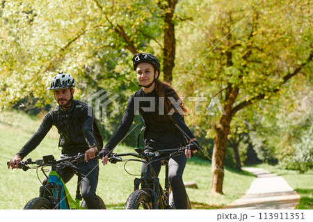 A blissful couple, adorned in professional cycling gear, enjoys a romantic bicycle ride through a park, surrounded by modern natural attractions, radiating love and happiness 113911351