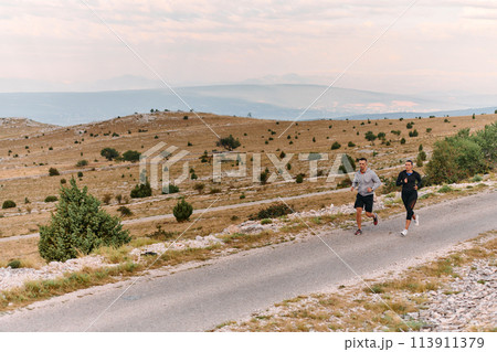 A couple dressed in sportswear runs along a scenic road during an early morning workout, enjoying the fresh air and maintaining a healthy lifestyle 113911379