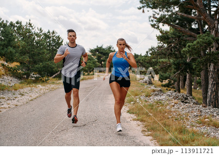 A couple dressed in sportswear runs along a scenic road during an early morning workout, enjoying the fresh air and maintaining a healthy lifestyle A couple dressed in sportswear runs along a scenic road during an early morning workout, enjoying the fresh air and maintaining a healthy lifestyle 113911721