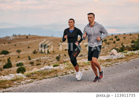 A couple dressed in sportswear runs along a scenic road during an early morning workout, enjoying the fresh air and maintaining a healthy lifestyle 113911735