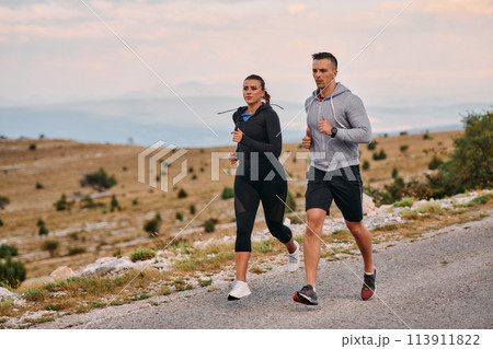 A couple dressed in sportswear runs along a scenic road during an early morning workout, enjoying the fresh air and maintaining a healthy lifestyle 113911822