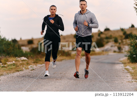 A couple dressed in sportswear runs along a scenic road during an early morning workout, enjoying the fresh air and maintaining a healthy lifestyle 113912204