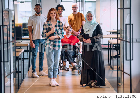 A diverse group of young business people walking a corridor in the glass-enclosed office of a modern startup, including a person in a wheelchair and a woman wearing a hijab 113912580