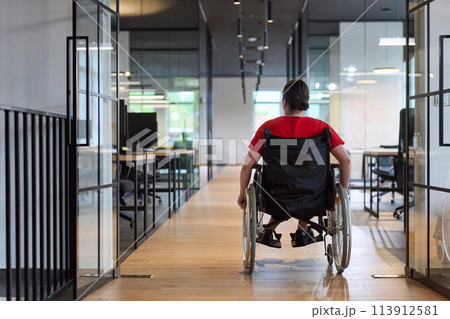 A modern young businesswoman in a wheelchair is surrounded by an inclusive workspace with glass-walled offices, embodying determination and innovation in the business world 113912581