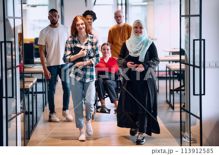 A diverse group of young business people walking a corridor in the glass-enclosed office of a modern startup, including a person in a wheelchair and a woman wearing a hijab A diverse group of young business people walking a corridor in the glass-enclosed office of a modern startup, including a person in a wheelchair and a woman wearing a hijab 113912585