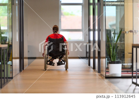 A modern young businesswoman in a wheelchair is surrounded by an inclusive workspace with glass-walled offices, embodying determination and innovation in the business world 113912645
