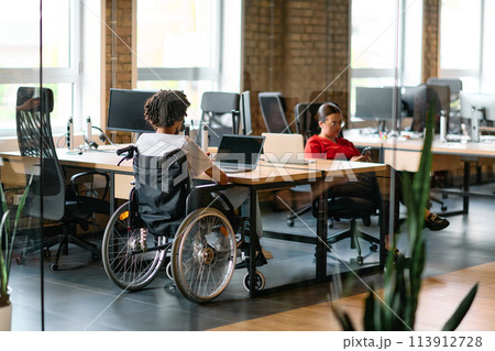 A young business group, including an African American businessman in a wheelchair, collaborates within a modern glass office, actively engaged around a computer and laptop, collectively solving 113912728