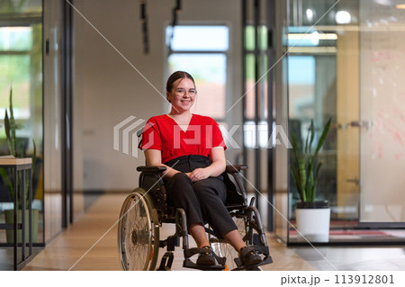 A modern young businesswoman in a wheelchair is surrounded by an inclusive workspace with glass-walled offices, embodying determination and innovation in the business world 113912801