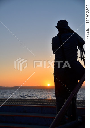 海に沈む日を見守る旅の記録者　黄昏の光をとらえて　夕日を追うカメラマン 113912802
