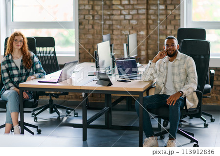 Group of colleagues, a woman with vibrant orange hair and a young African American businessman, sitting in a modern office space, symbolizing diverse collaboration and a dynamic work environment. 113912836