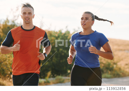 A couple dressed in sportswear runs along a scenic road during an early morning workout, enjoying the fresh air and maintaining a healthy lifestyle A couple dressed in sportswear runs along a scenic road during an early morning workout, enjoying the fresh air and maintaining a healthy lifestyle 113912856
