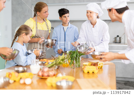 Friendly female chef in white uniform giving culinary lesson to tweens Friendly female chef in white uniform giving culinary lesson to tweens 113917842