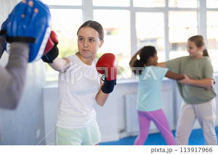 Focused girl hitting the focus mitts held by his boxing trainer in gym Focused girl hitting the focus mitts held by his boxing trainer in gym 113917966