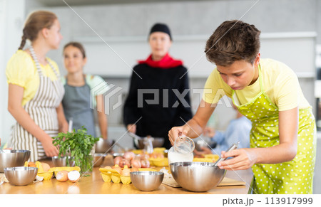 Cooking course - portrait of teenage boy in an apron who is learning to beat eggs to make pancakes at cooking class Cooking course - portrait of teenage boy in an apron who is learning to beat eggs to make pancakes at cooking class 113917999