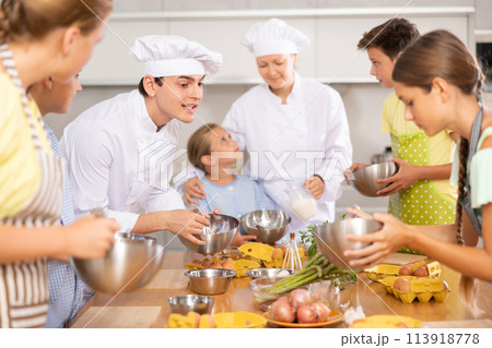 During cooking lesson, male cook tells children about culinary rules, woman assistant helps child During cooking lesson, male cook tells children about culinary rules, woman assistant helps child 113918778