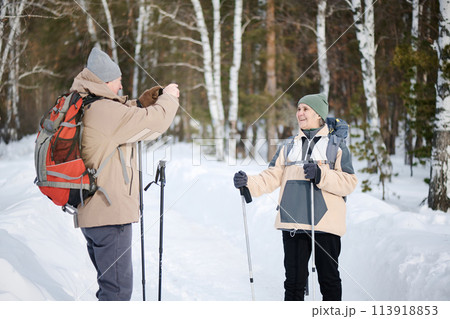Medium long shot of senior Caucasian tourist holding smartphone taking photo of his wife in forest park on winter day 113918853