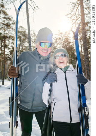 Vertical medium long shot of joyful active senior couple in winter sportswear and sunglasses standing outdoors posing for camera with skis and poles Vertical medium long shot of joyful active senior couple in winter sportswear and sunglasses standing outdoors posing for camera with skis and poles 113918895