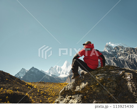 asian man sitting on top of rock looking at view with Mount Yangmaiyong (or Jampayang in Tibetan) in the distance in Yading, China 113925430