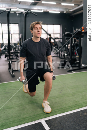 Vertical portrait of motivated young beginner athletic sportsman in activewear holding dumbbells doing squat lunges warm up training indoor at gym. Concept of healthy lifestyle, physical activity. Vertical portrait of motivated young beginner athletic sportsman in activewear holding dumbbells doing squat lunges warm up training indoor at gym. Concept of healthy lifestyle, physical activity. 113927042