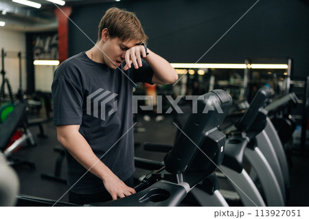 Portrait of tired beginner runner sportsman resting after running on treadmill during cardio session in gym. Satisfied fit male standing on treadmill in fitness gym, feeling exhausted after jogging. 113927051