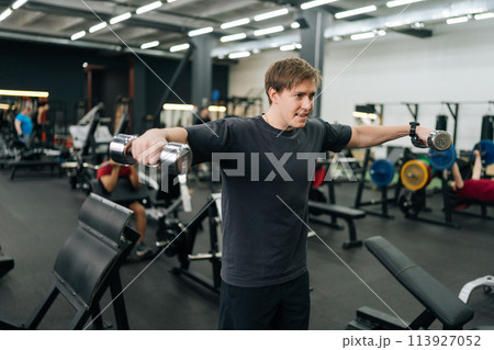 Portrait of motivated beginner sportsman hard time doing lateral arm raise exercise with dumbbells. Tired male athlete improves shoulders with dumbbell breeding. Concept of healthy lifestyle 113927052