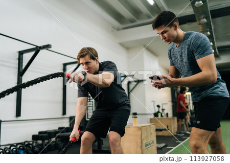 Portrait of dedicated sportsman exercising with battle rope while coach encouraging during cross training in health club standing with stopwatch. Concept of healthy lifestyle, physical sport activity. Portrait of dedicated sportsman exercising with battle rope while coach encouraging during cross training in health club standing with stopwatch. Concept of healthy lifestyle, physical sport activity. 113927058