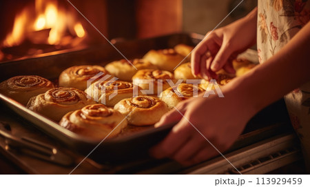 Close-up of a woman's hand taking warm, golden-brown rolls from a dimly lit oven in a cozy kitchen, evoking homemade comfort. 113929459